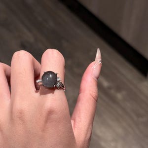 Puede incluir: Un anillo de plata con una piedra preciosa gris ovalada grande. El anillo tiene peque&ntilde;as piedras de acento transparentes a ambos lados de la piedra principal. El anillo se muestra en una mano con u&ntilde;as largas y cuidadas.