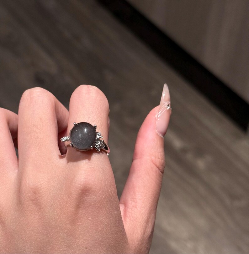 Puede incluir: Un anillo de plata con una piedra preciosa gris ovalada grande. El anillo tiene peque&ntilde;as piedras de acento transparentes a ambos lados de la piedra principal. El anillo se muestra en una mano con u&ntilde;as largas y cuidadas.