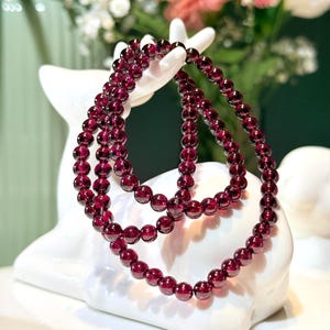 May include: A strand of deep red, translucent garnet beads arranged in a circular shape. The beads are polished, spherical, and uniform in size, reflecting light on their smooth surfaces. The necklace is draped over a white ceramic deer figurine.