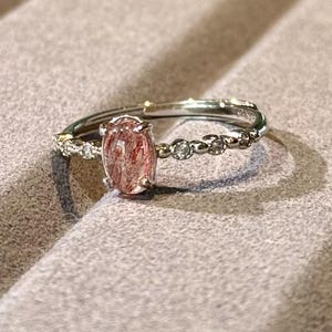 May include: A silver ring featuring a large, oval-shaped, reddish-brown gemstone. The band is adorned with small, clear gemstones and has an adjustable design. The ring is displayed on a gray surface.
