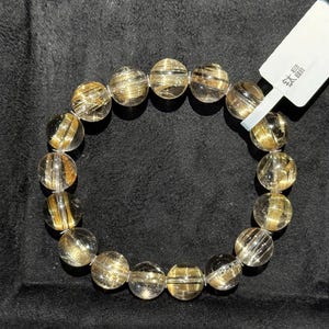 K&ouml;nnte beinhalten: Ein Armband aus klarem Quarz mit goldenen Rutil-Einschl&uuml;ssen. Die runden Perlen haben eine polierte Oberfl&auml;che, die einen funkelnden Effekt erzeugt. Das Armband wird auf einer schwarzen Oberfl&auml;che pr&auml;sentiert. Ein wei&szlig;es Etikett mit chinesischen Schriftzeichen ist angebracht.