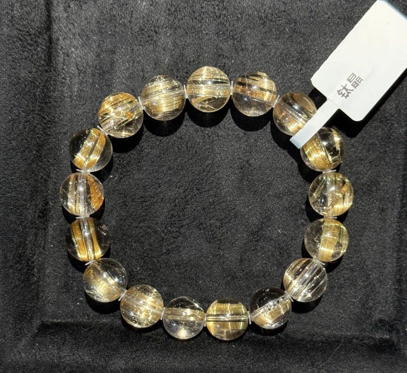 K&ouml;nnte beinhalten: Ein Armband aus klarem Quarz mit goldenen Rutil-Einschl&uuml;ssen. Die runden Perlen haben eine polierte Oberfl&auml;che, die einen funkelnden Effekt erzeugt. Das Armband wird auf einer schwarzen Oberfl&auml;che pr&auml;sentiert. Ein wei&szlig;es Etikett mit chinesischen Schriftzeichen ist angebracht.