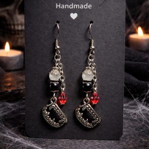 May include: A pair of handmade earrings on a black card. The earrings feature silver chains, white and black beads, red crystal beads, and silver vampire teeth charms. The card reads "Handmade" with a heart symbol.
