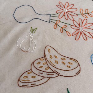 May include: Embroidered design on a cream-colored fabric featuring a vase with orange flowers, a garlic bulb, sliced bread, and cherries. The embroidery uses brown, orange, green, and blue thread.