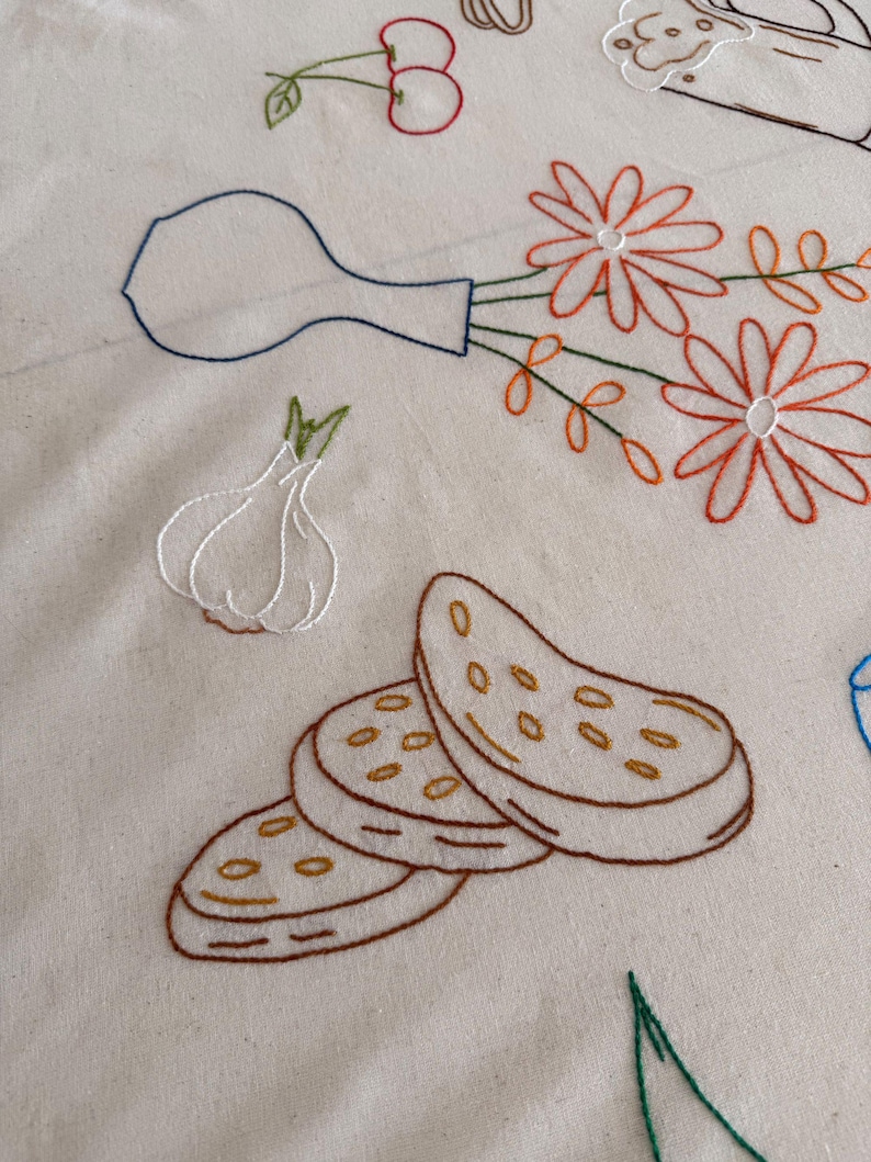 May include: Embroidered design on a cream-colored fabric featuring a vase with orange flowers, a garlic bulb, sliced bread, and cherries. The embroidery uses brown, orange, green, and blue thread.