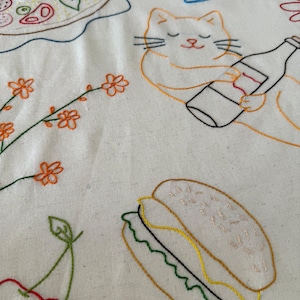 May include: Embroidered fabric featuring food and drink designs. Includes a pizza, a wine glass, a cat holding a bottle, a sandwich, cherries, and floral elements. The embroidery uses various colors on a white background.