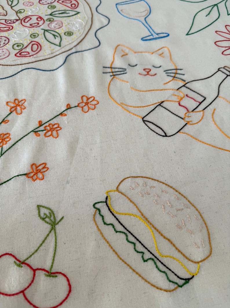 May include: Embroidered fabric featuring food and drink designs. Includes a pizza, a wine glass, a cat holding a bottle, a sandwich, cherries, and floral elements. The embroidery uses various colors on a white background.