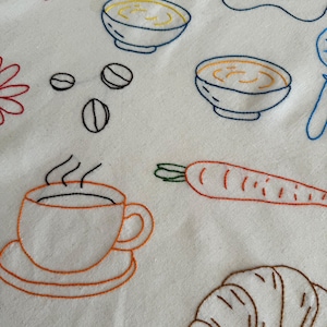 May include: Embroidered white fabric featuring various food items. Includes a coffee cup, croissant, carrot, bowls of soup, coffee beans, and a strawberry. The designs are outlined in orange, brown, blue, and red thread, creating a charming, handmade aesthetic.