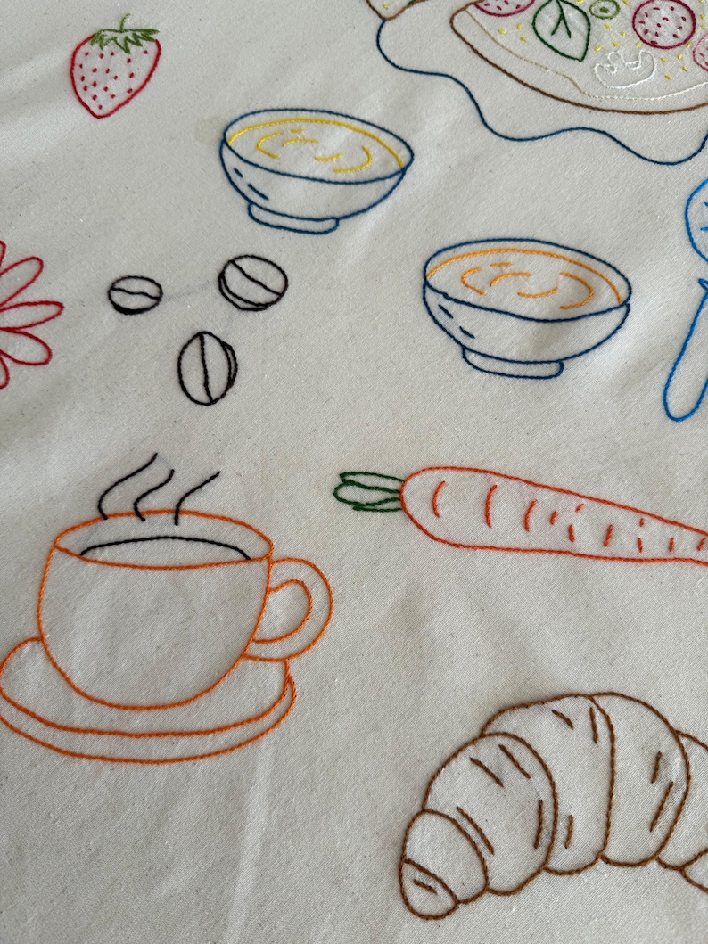 May include: Embroidered white fabric featuring various food items. Includes a coffee cup, croissant, carrot, bowls of soup, coffee beans, and a strawberry. The designs are outlined in orange, brown, blue, and red thread, creating a charming, handmade aesthetic.