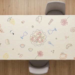 May include: Beige rectangular tablecloth with colorful line drawings of food and drink items, including pizza, wine glasses, flowers, and a dog holding a bottle. The tablecloth is set on a wooden table with four gray chairs.
