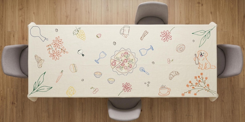May include: Beige rectangular tablecloth with colorful line drawings of food and drink items, including pizza, wine glasses, flowers, and a dog holding a bottle. The tablecloth is set on a wooden table with four gray chairs.