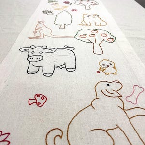 Puede incluir: Camino de mesa color crema con diseños bordados de animales y plantas. Presenta una vaca, un perro, un canguro, árboles, flores y otras ilustraciones caprichosas en hilo marrón, negro, verde y rojo. El camino de mesa está colocado sobre una mesa blanca.