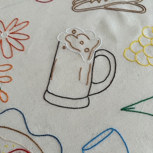 May include: Embroidered fabric with food and drink designs. Features a beer mug with foam, cherries, a baguette, a flower, grapes, a slice of pizza, a wine glass, and a green leaf. The designs are outlined in various colors.