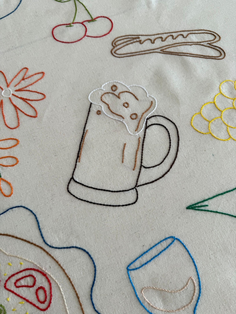 May include: Embroidered fabric with food and drink designs. Features a beer mug with foam, cherries, a baguette, a flower, grapes, a slice of pizza, a wine glass, and a green leaf. The designs are outlined in various colors.
