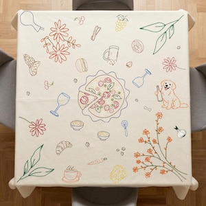 May include: A square, off-white tablecloth with embroidered illustrations of food and drink items. The design includes a pizza, wine glasses, flowers, and a dog holding a bottle. The tablecloth is set on a square table with two gray chairs.