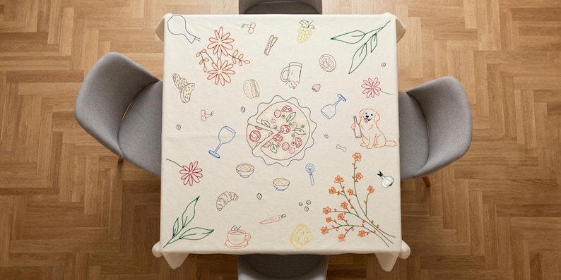 May include: A square, off-white tablecloth with embroidered illustrations of food and drink items. The design includes a pizza, wine glasses, flowers, and a dog holding a bottle. The tablecloth is set on a square table with two gray chairs.