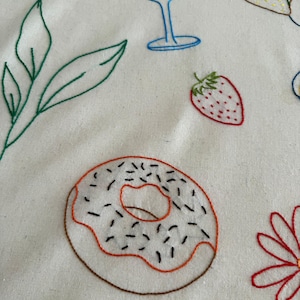 May include: Embroidered white fabric featuring colorful food and drink designs. A donut with sprinkles, a strawberry, a wine glass, and other food items are stitched in various colors. The design has a handmade, whimsical aesthetic.