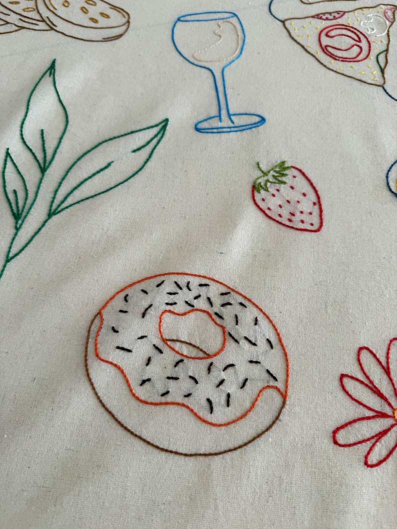 May include: Embroidered white fabric featuring colorful food and drink designs. A donut with sprinkles, a strawberry, a wine glass, and other food items are stitched in various colors. The design has a handmade, whimsical aesthetic.