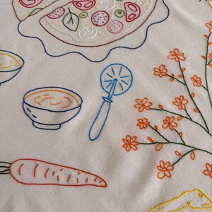 May include: Embroidered white fabric featuring a pizza with a slice cut out, a bowl of soup, a carrot, a pizza cutter, a cat, a sprig of orange flowers, and a piece of cheese. The designs are outlined in various colors.