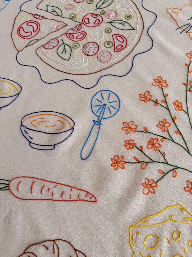 May include: Embroidered white fabric featuring a pizza with a slice cut out, a bowl of soup, a carrot, a pizza cutter, a cat, a sprig of orange flowers, and a piece of cheese. The designs are outlined in various colors.