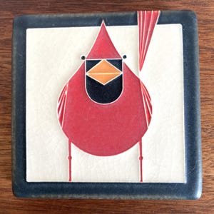 May include: A square ceramic tile featuring a stylized red cardinal bird graphic centered on a cream-colored background with a crackle glaze finish. The bird has a black face mask, an orange beak, and is framed by a dark charcoal border.
