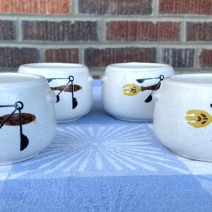 May include: Four white ceramic bowls with speckled surfaces and small handles. Each bowl features a design of a yellow fork, brown spoon, and black utensils. The bowls are arranged on a blue and white checkered tablecloth.