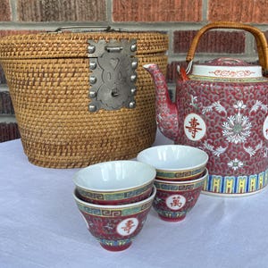 May include: A woven rattan basket with a metal latch, a red porcelain teapot with white floral patterns and Chinese characters, and three matching porcelain teacups featuring red, yellow, and blue decorative borders and Chinese calligraphy.