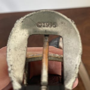 Pu&ograve; includere: Primo piano di una fibbia argentata con una cornice rettangolare e una barra centrale. La fibbia ha un aspetto usurato e l'anno "&copy;1995" &egrave; stampato sulla parte superiore. La fibbia &egrave; attaccata a una cintura di cuoio marrone.