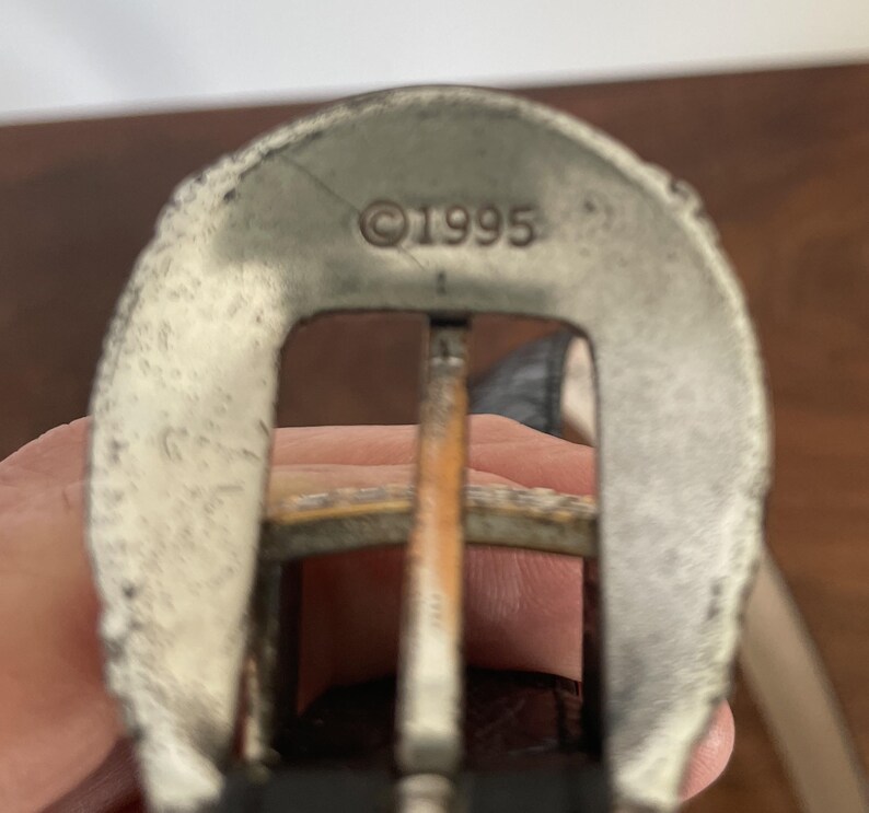 Pu&ograve; includere: Primo piano di una fibbia argentata con una cornice rettangolare e una barra centrale. La fibbia ha un aspetto usurato e l'anno "&copy;1995" &egrave; stampato sulla parte superiore. La fibbia &egrave; attaccata a una cintura di cuoio marrone.