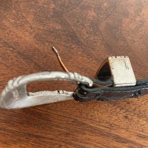 Pu&ograve; includere: Primo piano di una fibbia argentata con un design decorativo. La fibbia &egrave; attaccata a una cintura di pelle nera. La fibbia &egrave; su una superficie di legno.