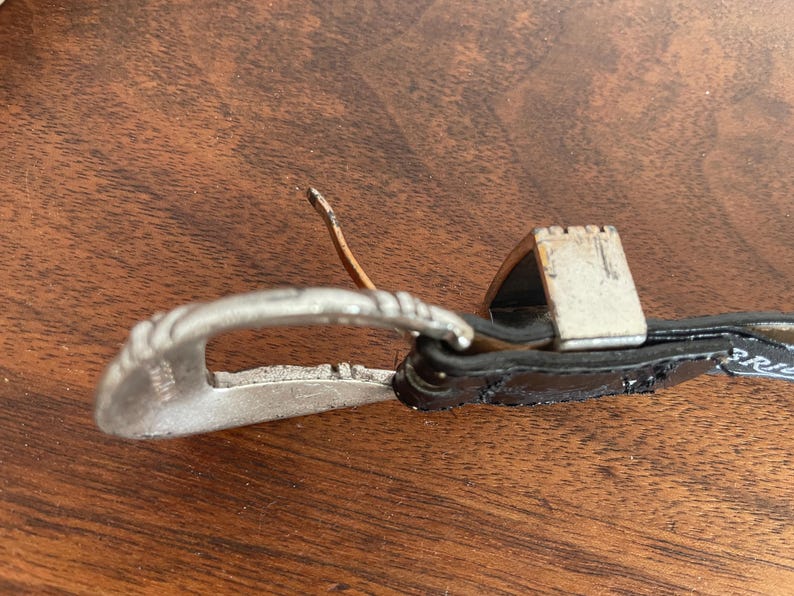 Pu&ograve; includere: Primo piano di una fibbia argentata con un design decorativo. La fibbia &egrave; attaccata a una cintura di pelle nera. La fibbia &egrave; su una superficie di legno.