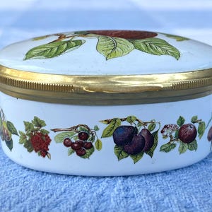 May include: A white, oval trinket box with a gold-colored rim and lid. The lid features a large red fruit with green leaves. The sides are decorated with illustrations of various fruits, including cherries and plums.