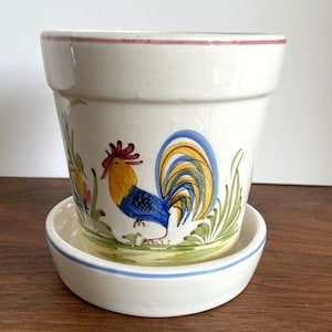 May include: A white ceramic flower pot with a matching saucer. The pot features a hand-painted rooster design in blue, yellow, and red. The rim of the pot has a thin pink stripe, and the saucer has a blue stripe.