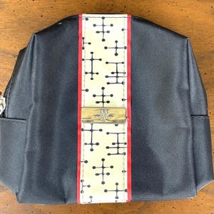 May include: Black cosmetic bag with a central cream-colored panel featuring a repeating black atomic design. The panel is bordered by red stripes and a gold-colored metal clasp. A silver zipper is visible on the side.