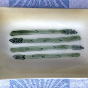 May include: A rectangular ceramic dish with a cream-colored surface. Four green asparagus spears are painted on the dish. The dish is set on a blue and white checkered tablecloth.