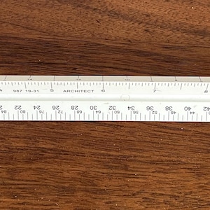 May include: A white architectural scale ruler with multiple measurement scales. The ruler is made of plastic and is marked with both inch and foot measurements. The ruler is approximately 12 inches long.