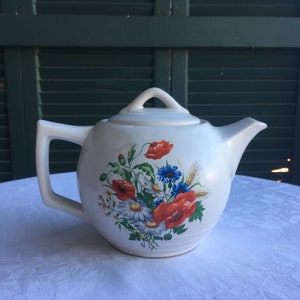 May include: Cream-colored ceramic teapot with a floral design featuring red poppies, blue cornflowers, and white daisies. The teapot has a handle, a spout, and a lid with a loop. The teapot is sitting on a white tablecloth.