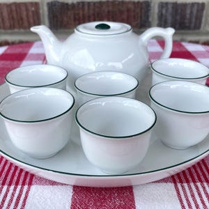May include: A white porcelain tea set with a teapot, six cups, and a serving tray. The cups and tray have a green trim. The teapot has a green knob on the lid. The set is arranged on a red and white checkered tablecloth.