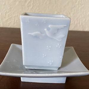 May include: A light blue ceramic teacup and saucer set with a raised design of a bird in flight. The set has a delicate, elegant look.