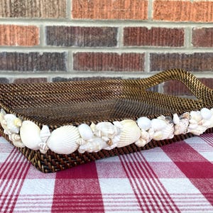 May include: A rectangular woven brown basket with a handle, decorated with white seashells along the rim. The basket sits on a red and white checkered tablecloth, with a brick wall in the background.