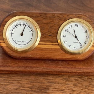 May include: A wooden desk clock with a thermometer and a clock face. The thermometer displays Fahrenheit, and the clock has gold-colored trim. The clock face reads "Quartz". The wooden base is a dark brown color.