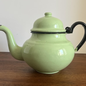May include: A light green enamel teapot with a black handle and rim. The teapot has a rounded body, a curved spout, and a lid with a small knob. The teapot is sitting on a wooden surface.