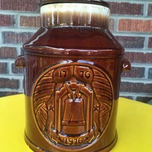May include: Brown ceramic cookie jar shaped like a milk jug with a lid. The jar has a design of a bell with the years 1776 and 1976 on it.