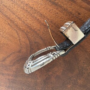 Pu&ograve; includere: Primo piano di una cintura in pelle nera con fibbia e passante argentati. La fibbia &egrave; rettangolare con un design strutturato. Il passante &egrave; anch'esso argentato e presenta un motivo decorativo.