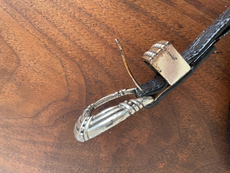 Pu&ograve; includere: Primo piano di una cintura in pelle nera con fibbia e passante argentati. La fibbia &egrave; rettangolare con un design strutturato. Il passante &egrave; anch'esso argentato e presenta un motivo decorativo.