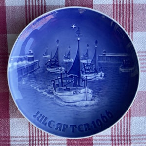 May include: Blue and white Christmas plate with a scene of fishing boats sailing into a harbor at night. The plate has the text "Julgaften 1966" on the bottom.