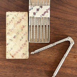 May include: An antique nutcracker and a set of silver-toned tools in a decorative box. The box and lid have a floral pattern. The tools are arranged in a row, and the nutcracker is open, ready for use. The items are on a wooden surface.