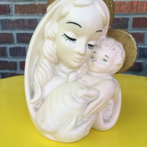 May include: A white ceramic figurine of the Virgin Mary holding a baby Jesus. The figurine is painted with gold accents and has a halo around Mary's head.