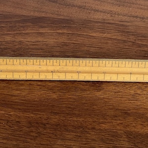 May include: A wooden architectural scale ruler, marked with imperial measurements. The ruler is light brown and has multiple scales for architectural drawings. The scale is approximately 12 inches long.