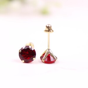 May include: A pair of gold stud earrings with red gemstones. The earrings are on a white surface.